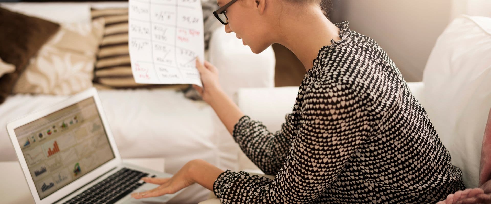 Pessoa analisando dados financeiros em um laptop no quarto, com gráficos e relatórios em papel na mão, demonstrando análise financeira pessoal.