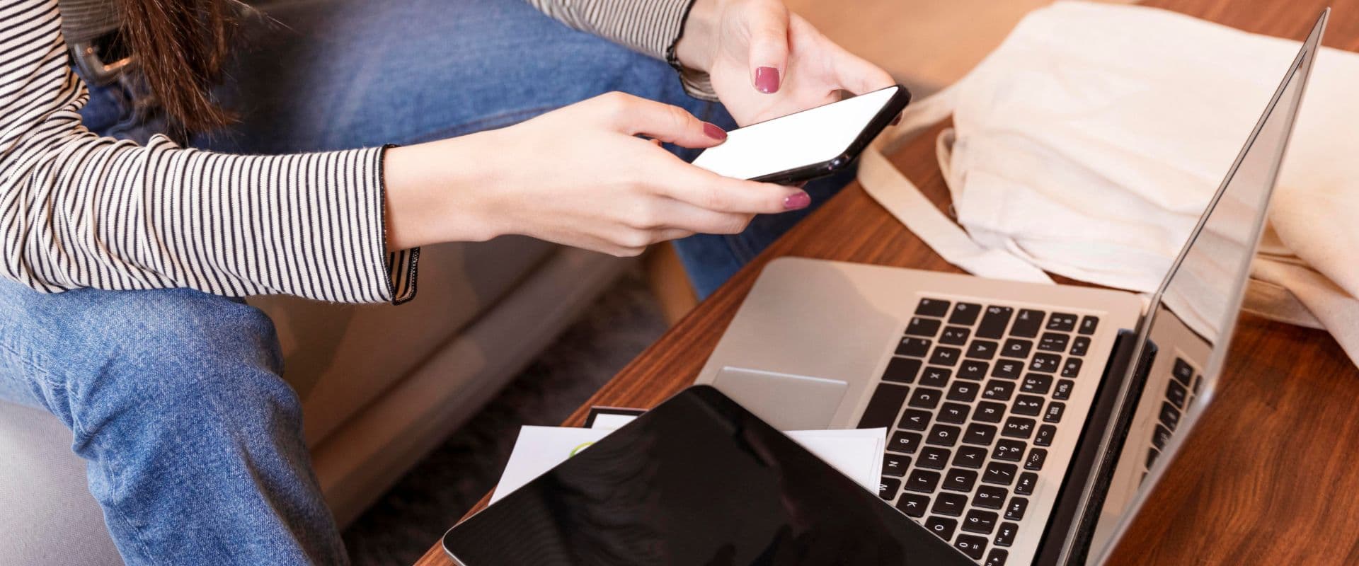 Pessoa usando smartphone perto de laptop e tablet em uma mesa de trabalho, com papéis ao redor, ambiente de trabalho moderno.
