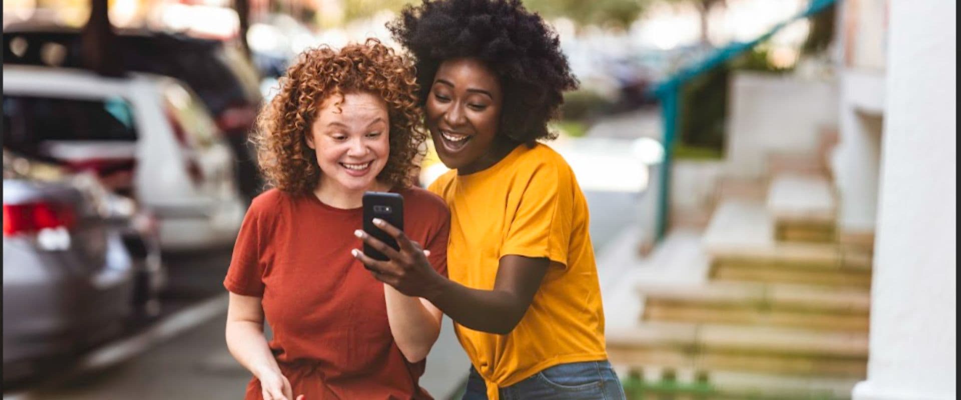 Duas mulheres felizes olhando para um celular, uma delas com cabelo cacheado e blusa vermelha e outra com cabelo afro e moletom amarelo, em ambiente externo, demonstrando conexão e alegria.