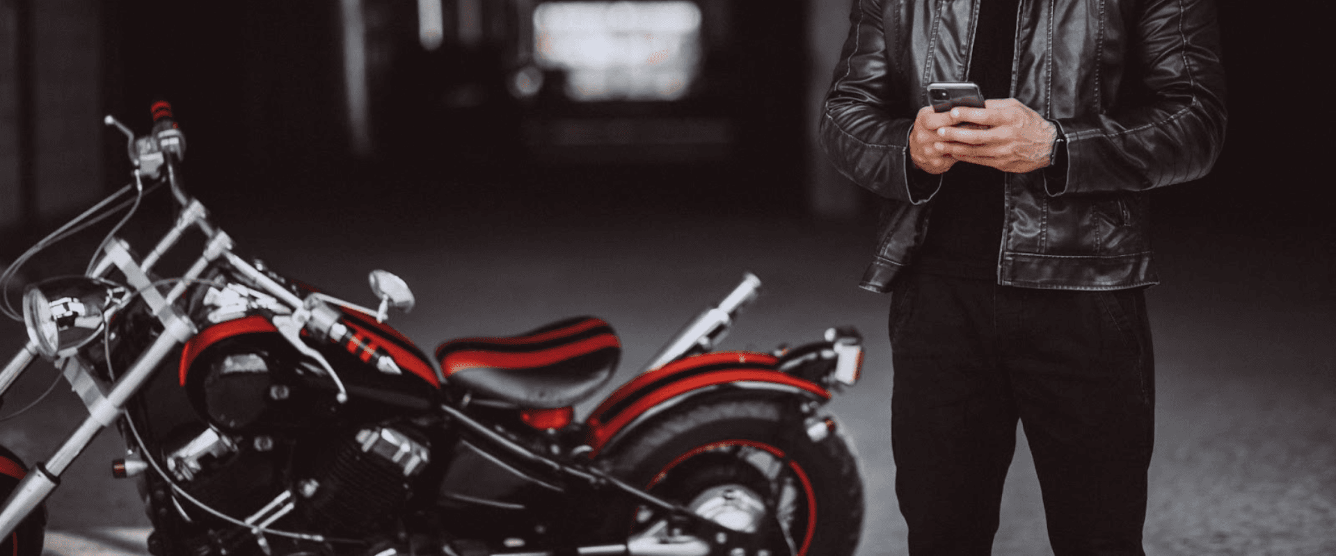 Homem com jaqueta de couro usando celular ao lado de uma motocicleta clássica preta e vermelha, sob luzes de uma garagem moderna.