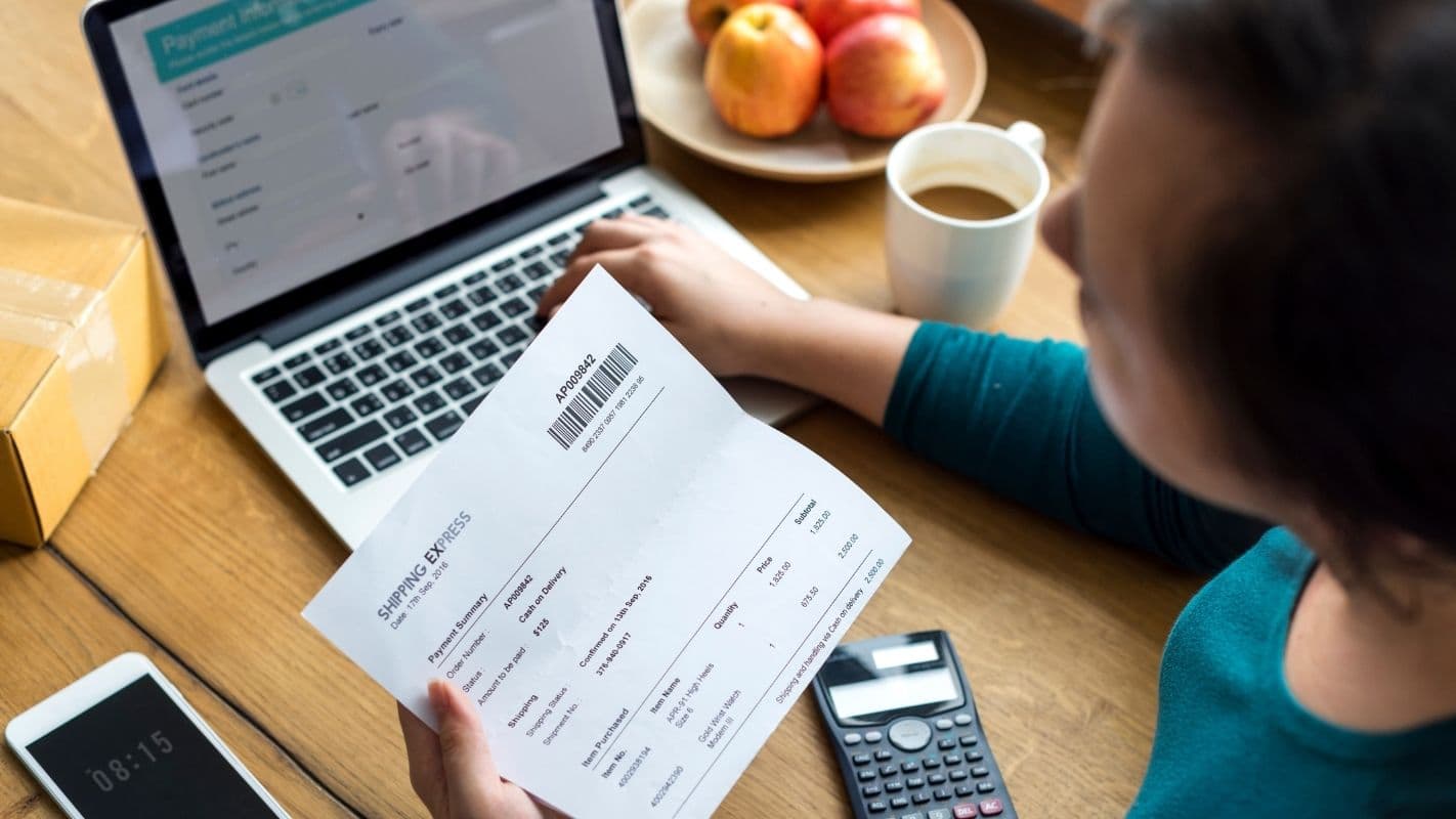 Pessoa pagando uma fatura enquanto trabalha com laptop, smartphone e calculadora na mesa de escritório.
