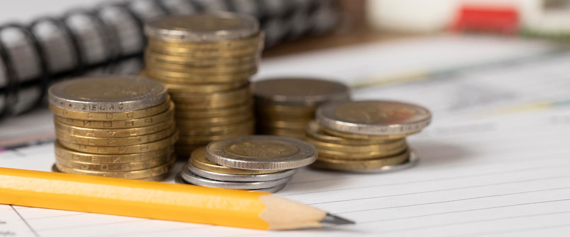 Pilhas de moedas de diferentes valores empilhadas ao lado de um caderno e um lápis amarelo, simbolizando economia e finanças pessoais.