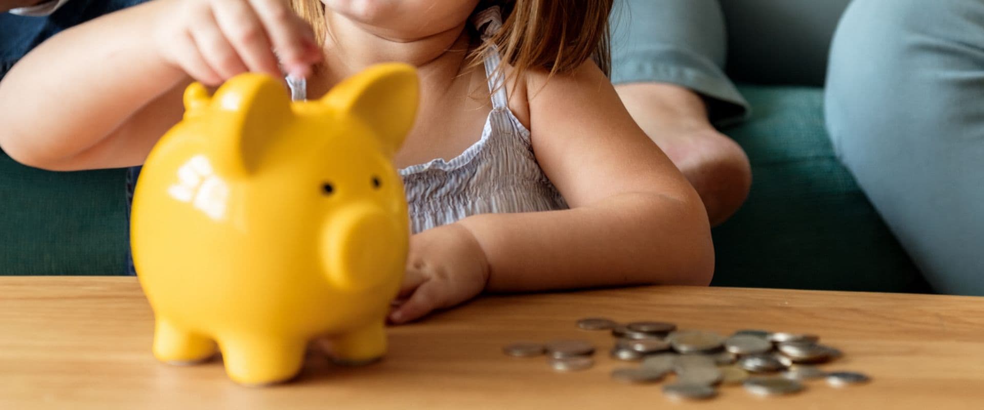 Criança colocando uma moeda na boca do cofrinho de porquinho amarelo, simbolizando economia e finanças para crianças.