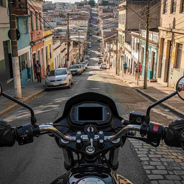 Visão de uma rua inclinada na cidade, vista do ponto de vista de uma motocicleta, com carros estacionados e edifícios coloridos ao redor.