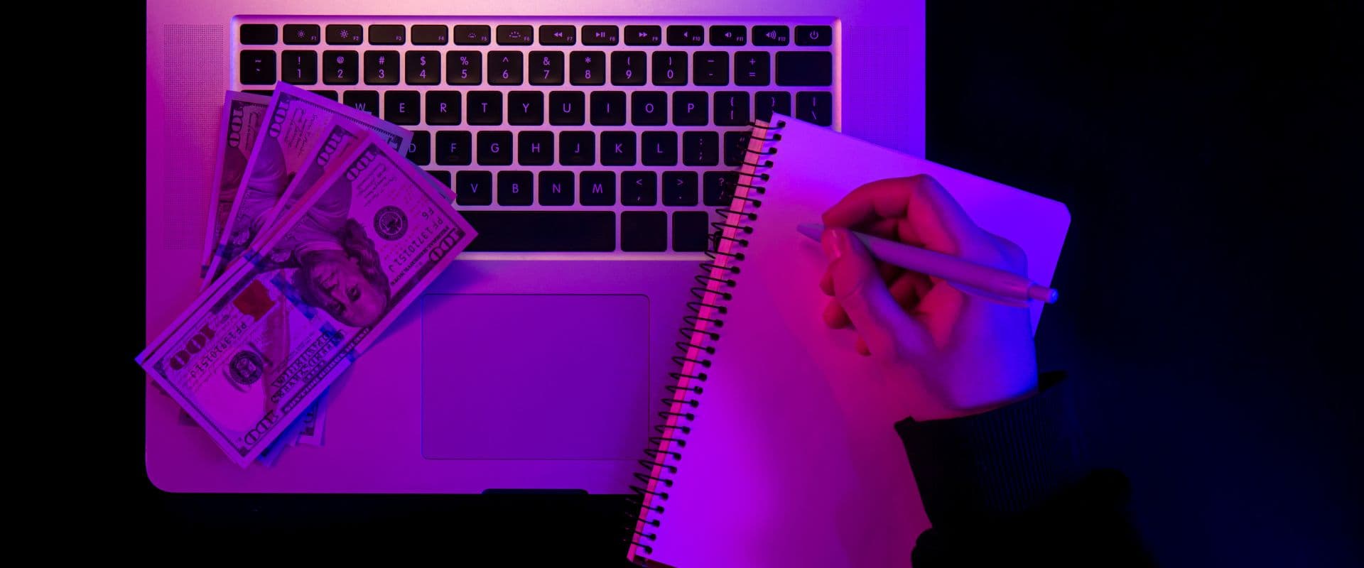 Imagem de um laptop com teclado iluminado, pilhas de dinheiro e uma mão escrevendo em um caderno, todos com iluminação roxa e azul, simbolizando negócios, investimentos e ganhos financeiros.