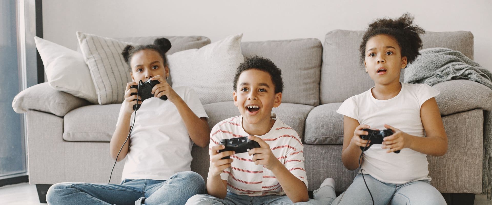 Três crianças se divertindo e jogando videogame juntas na sala de estar, sentadas no chão em frente a um sofá cinza. Elas parecem estar em um momento de diversão e interação.