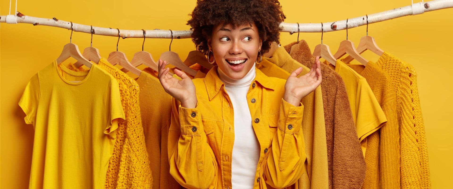Mulher sorridente de cabelo cacheado fazendo escolhas de roupas amarelas em uma loja de moda, promovendo looks fashion e tendências coloridas.