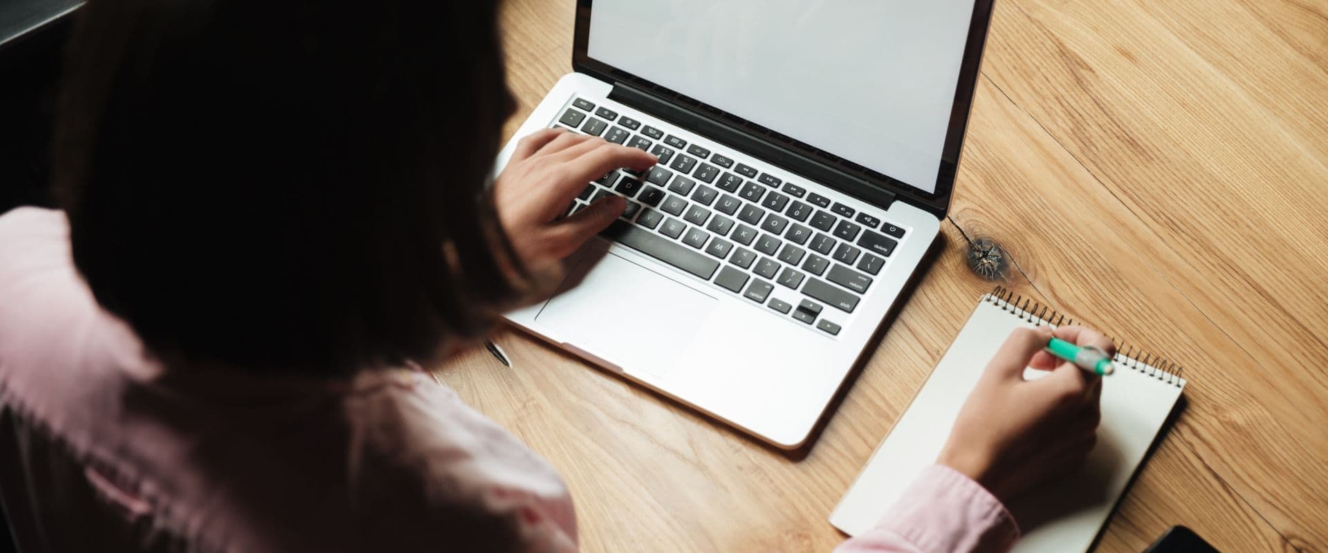 Pessoa trabalhando no computador portátil com caderno e caneta sobre uma mesa de madeira, ambiente de estudo ou trabalho remoto.