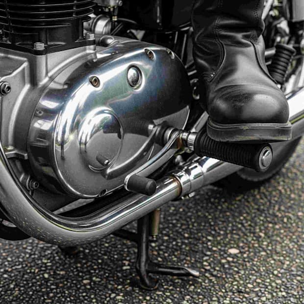 Close-up da engrenagem e pedal de uma motocicleta, além de uma bota de couro do motociclista, destacando detalhes do motor e da parte de condução.