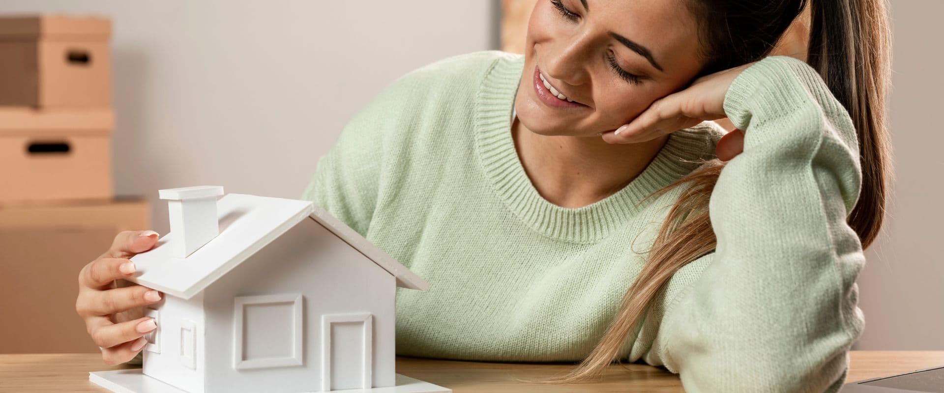 Mulher sorrindo observando uma maquete de uma casa, simbolizando sonhos de comprar ou construir uma casa, representação de planejamento imobiliário ou sonho da casa própria.
