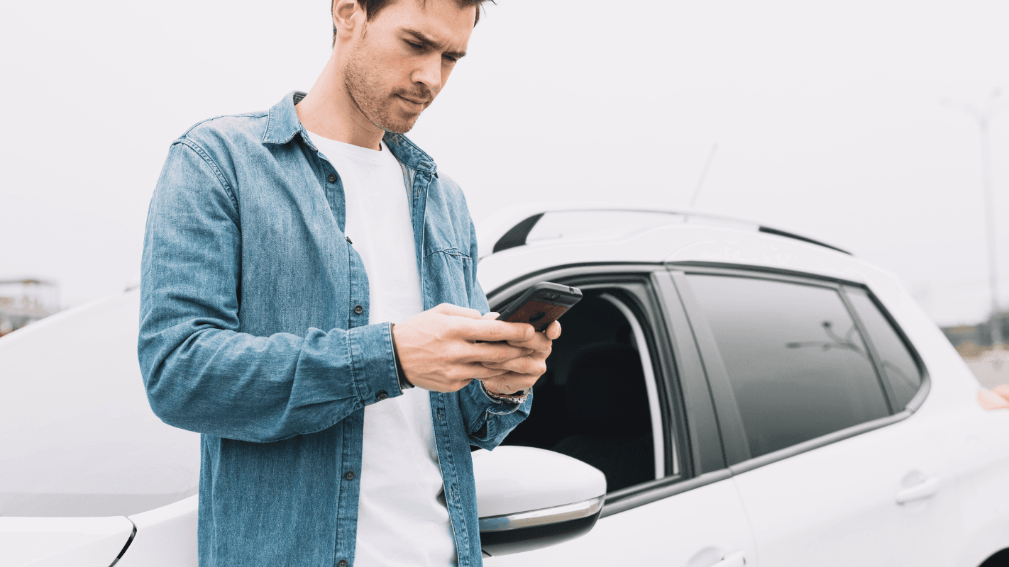 Homem usando celular ao lado de um carro branco em um estacionamento, realizando uma ligação ou enviando mensagem.