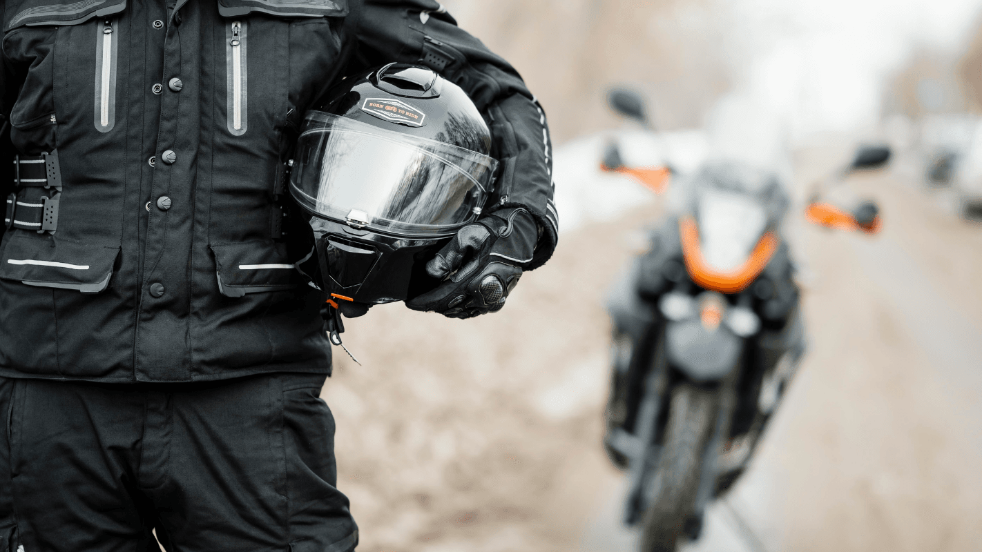 Homem com roupa de proteção para motocicleta segurando capacete preto, ao lado de moto estilo adventure, em ambiente ao ar livre.