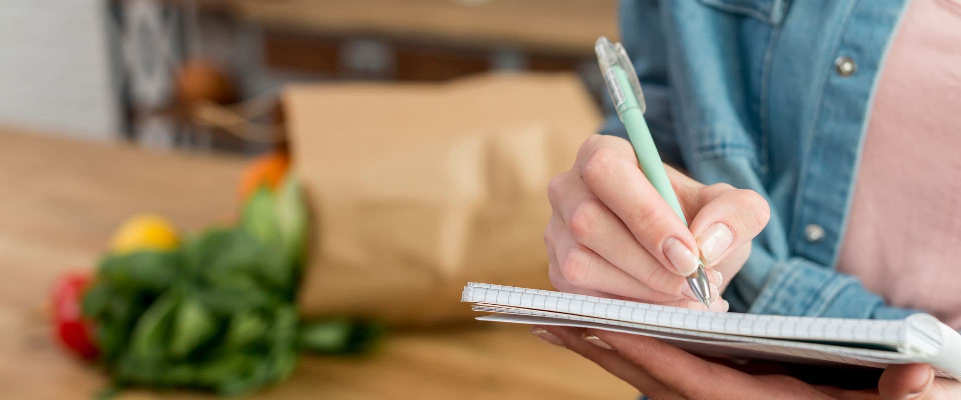 Pessoa anotando em um caderno com vegetais no fundo, provavelmente planejando refeições ou listando compras. Ambiente de cozinha ou mercado.