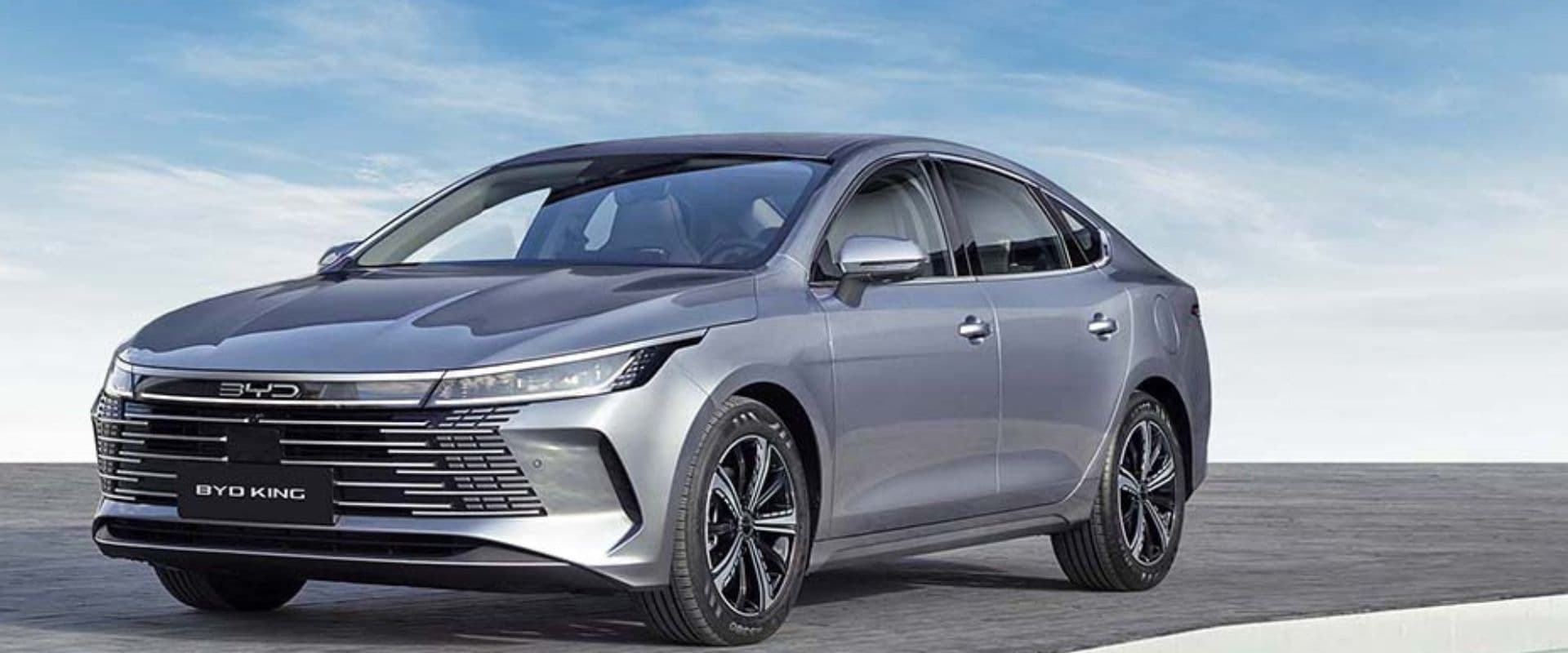 Imagem de um carro BYD King, sedan moderno e elegante de cor cinza, com design aerodinâmico e detalhes tecnológicos, estacionado ao ar livre sob um céu azul.