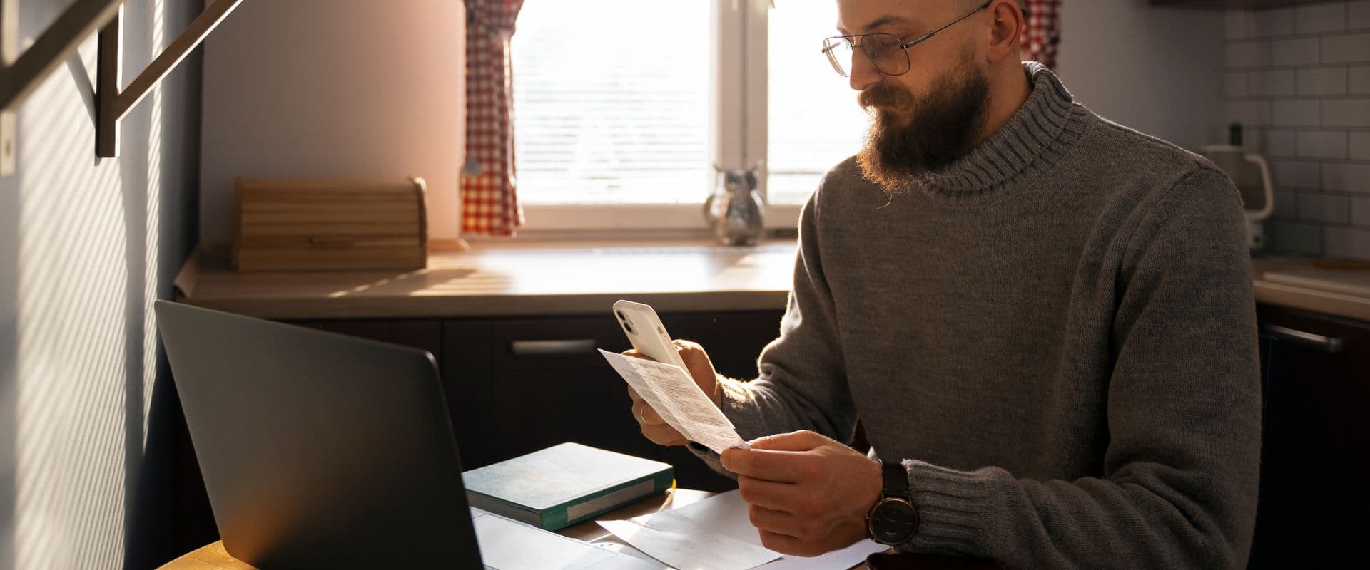 Homem com barba usando óculos e suéter cinza trabalha com notebook e celular em uma cozinha moderna bem iluminada.