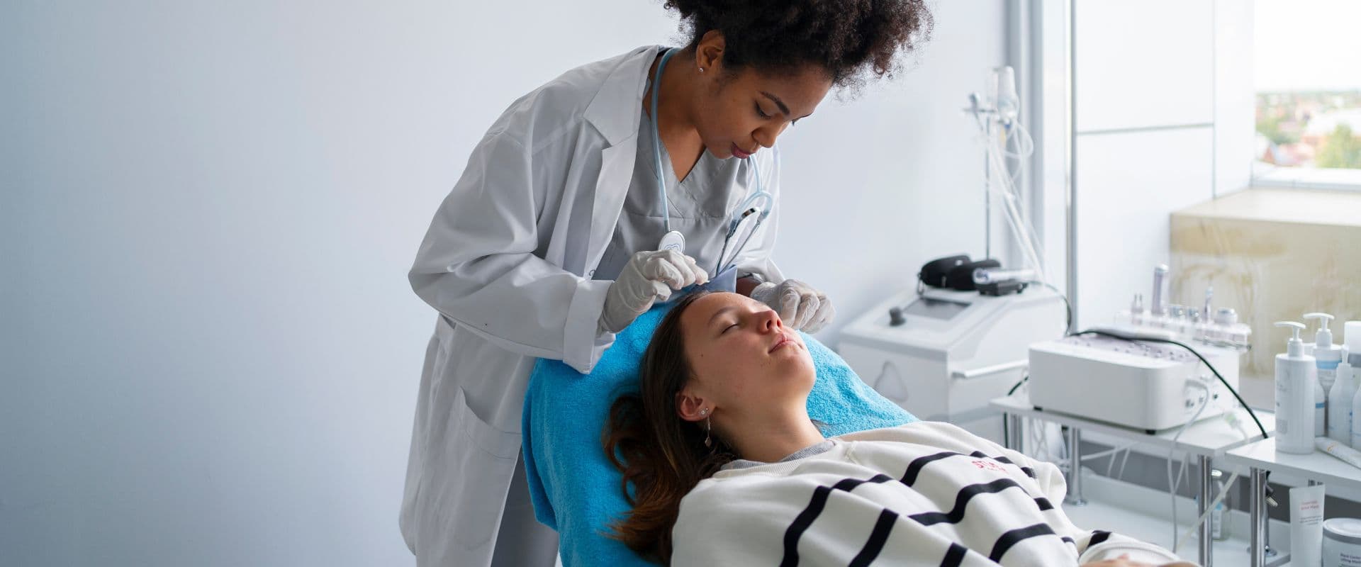 Profissional de saúde realizando procedimento estético facial em paciente deitada em clínica de estética com equipamentos modernos ao fundo.
