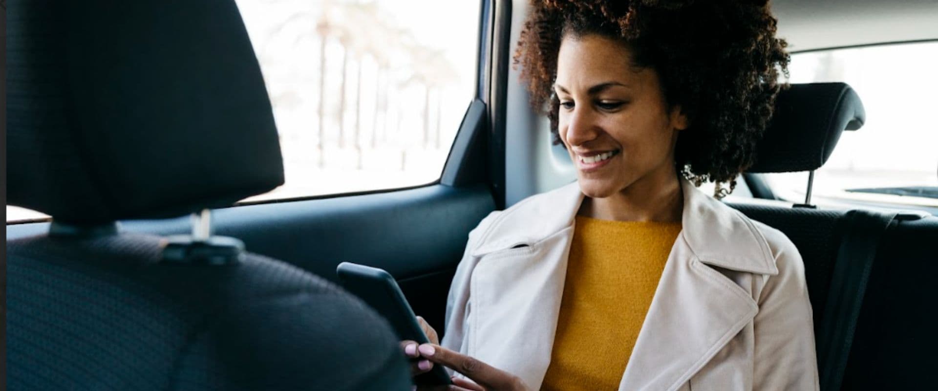 Mulher sorridente usando smartphone no banco de trás de um carro, desfrutando de um momento agradável enquanto viaja, representando segurança e bem-estar.