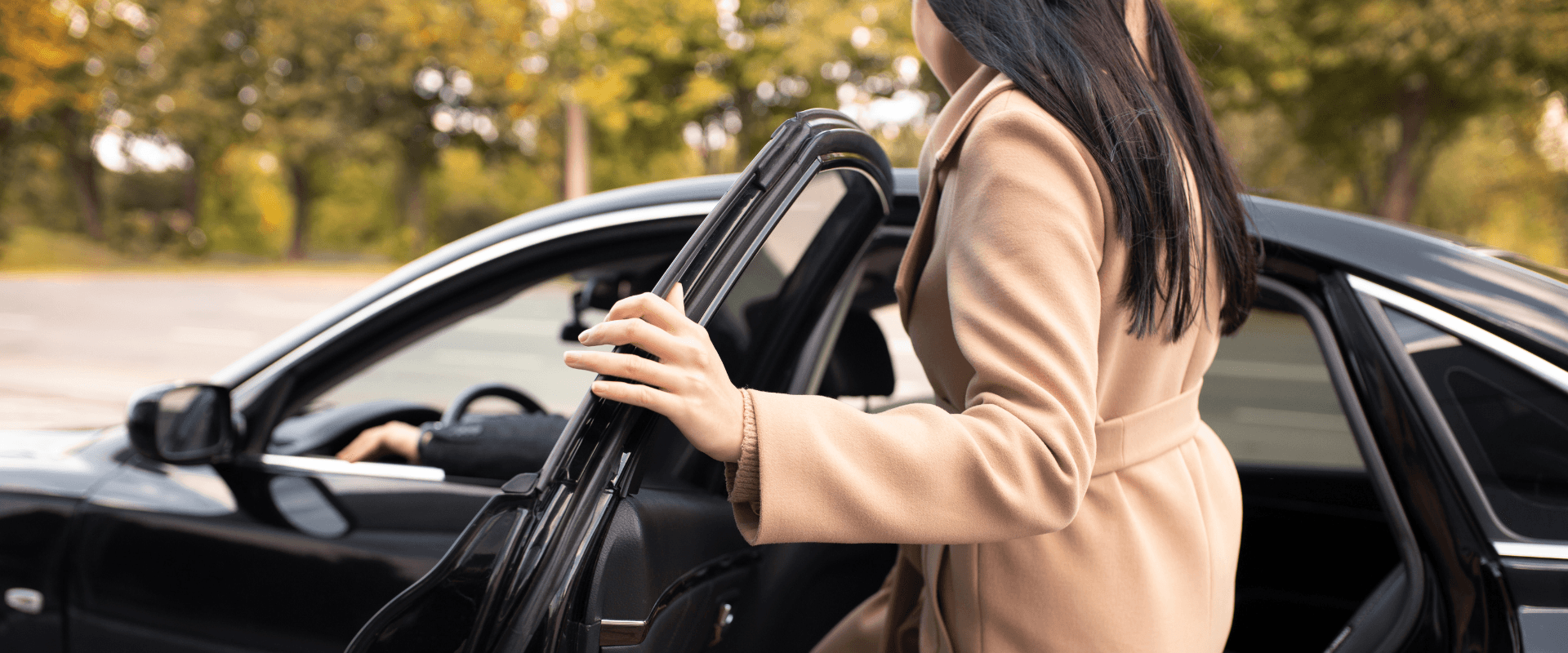 Mulher entrando em um carro preto durante o dia, com árvores ao fundo, em uma cena urbana ou rural. Carro parado.