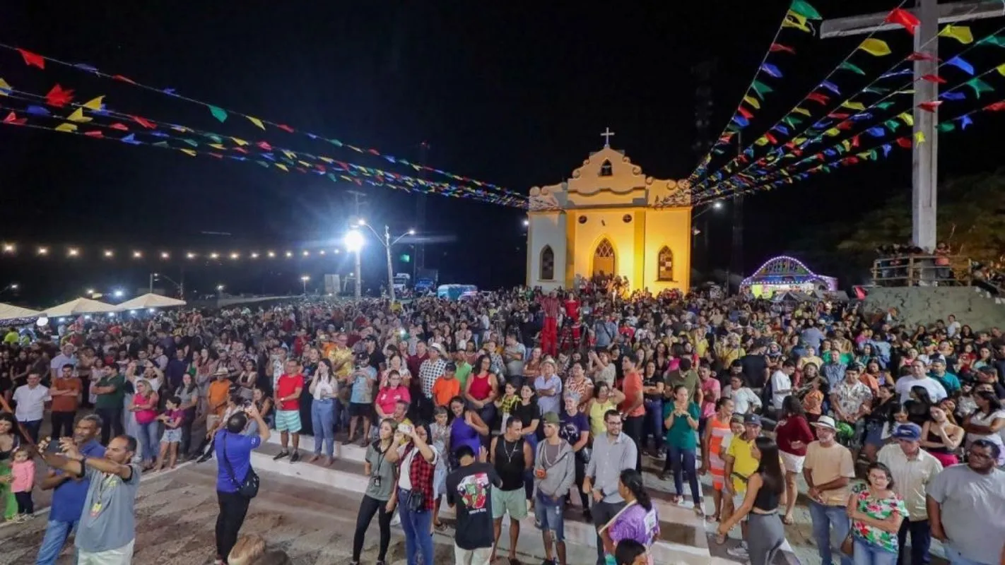 Grande evento religioso ao ar livre à noite, com uma multidão de fiéis e uma igreja iluminada ao fundo decorada com bandeirinhas coloridas.