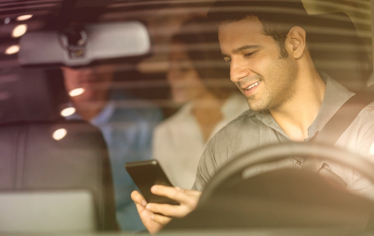 Motorista mexe no celular; no banco traseiro do carro, está um casal de passageiros - 6 vantagens para trabalhar com aplicativos de transporte