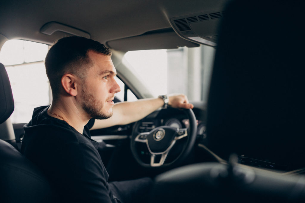 Na imagem há uma pessoa dentro de um carro, segurando com uma das mãos o volante, enquanto reflete como a renovação simplificada da CNH é rápida.