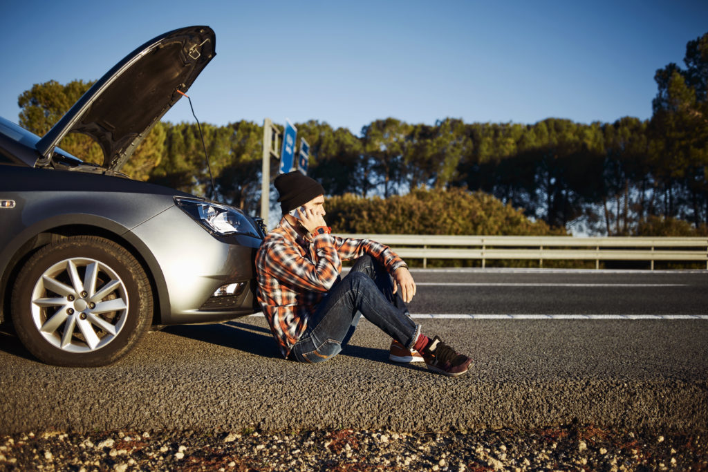 Na imagem há uma pessoa caucasiana sentada na frente de um carro parado, lidando com as uma das consequências da gasolina adulterada. o não funcionamento correto do motor.