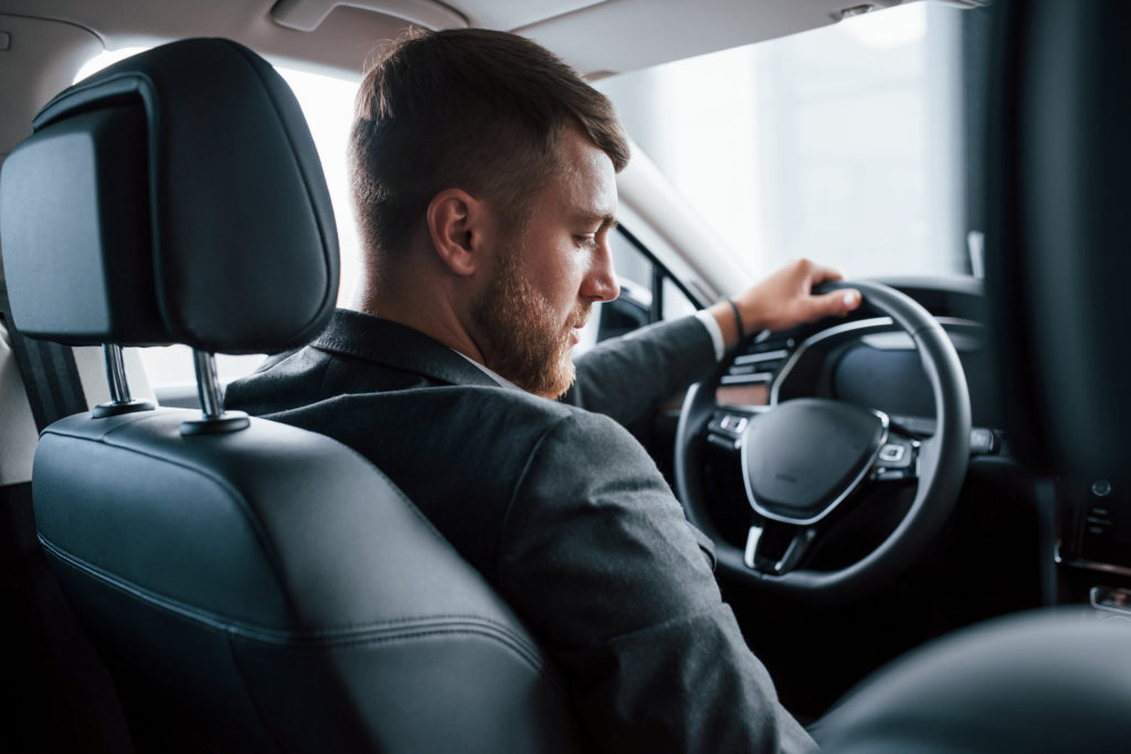 Na imagem há um homem caucasiano, segurando com mão esquerda o volante e olhando para a região do câmbio do carro.