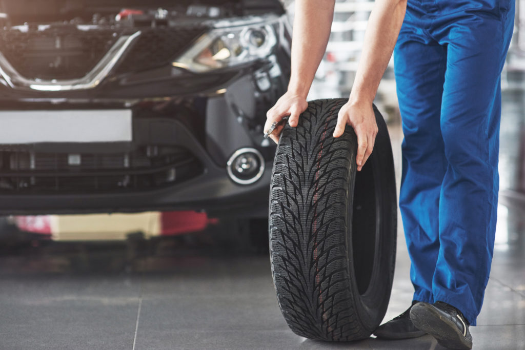 Na imagem há uma pessoa segurando um pneu próximo a um carro preto, possivelmente trocando para evitar aquaplanagem.