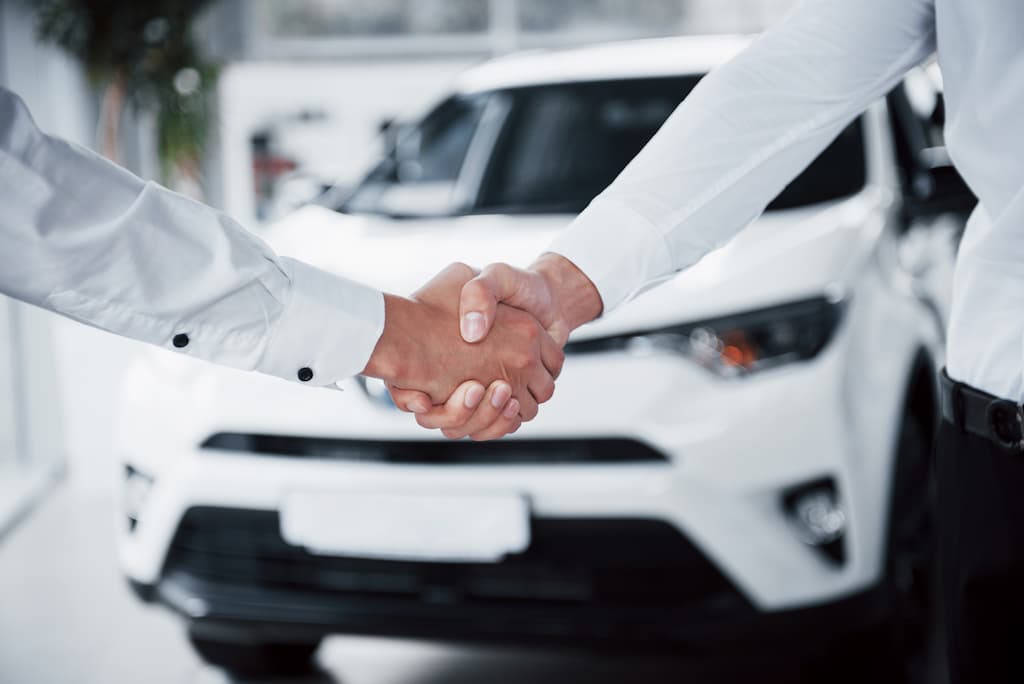 Na imagem há duas pessoas apertando aos mãos em frente a um carro branco.