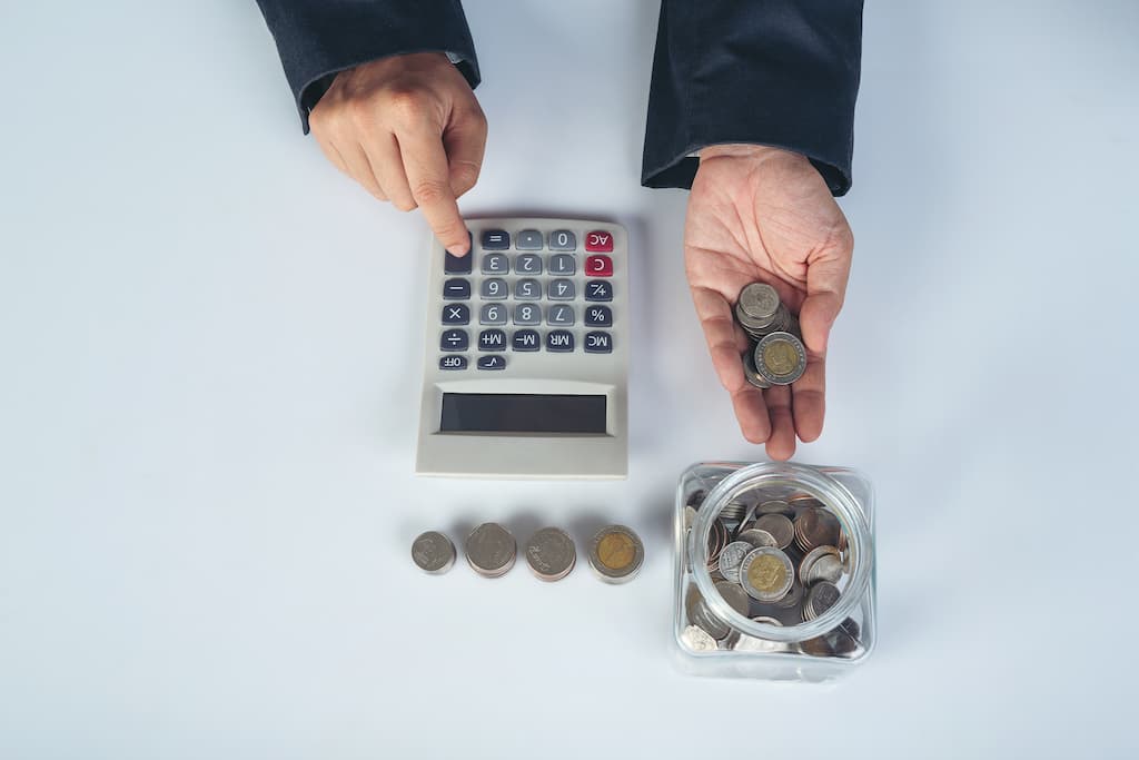 Na imagem há uma pessoa mexendo em uma calculadora, enquanto segura com a outra mão algumas moedas.
