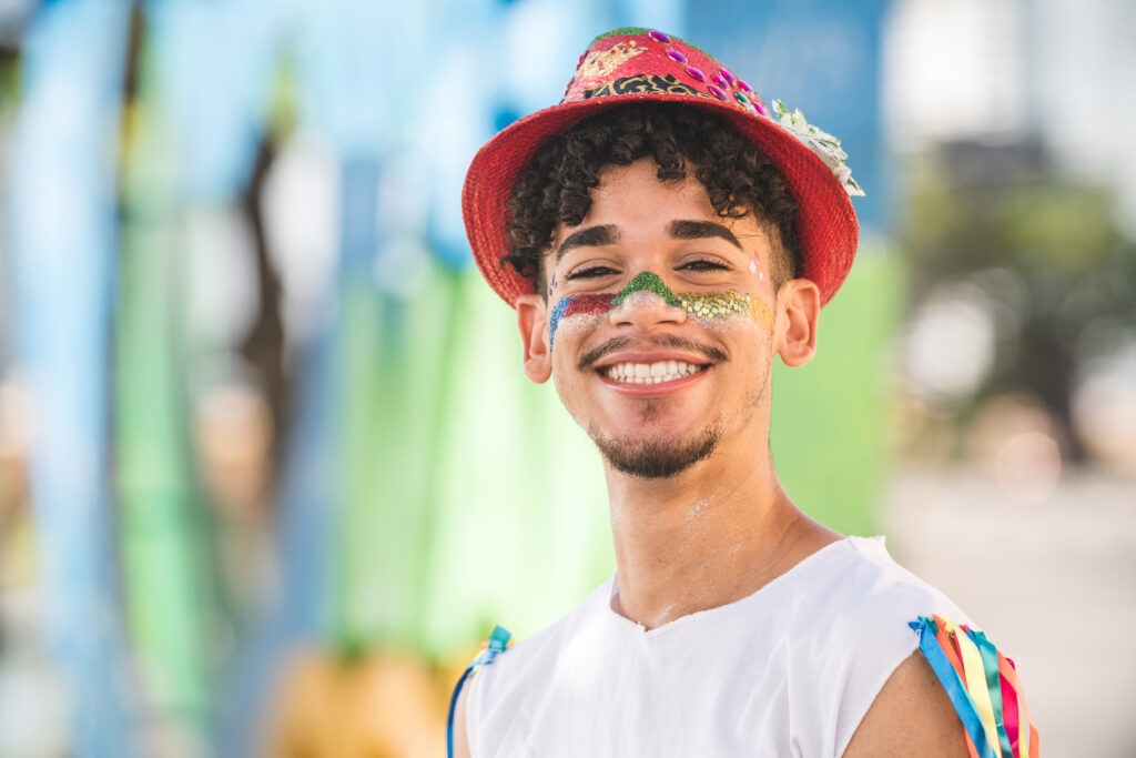Na imagem há uma pessoa do gênero masculino que está sorrindo para a foto e vestido com roupas temáticas de carnaval.