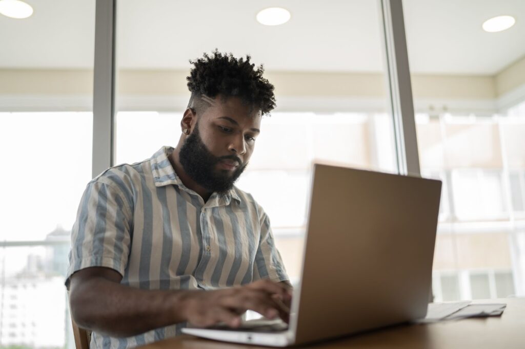 Na imagem há um homem negro mexendo em um computador apoiado em uma mesa.